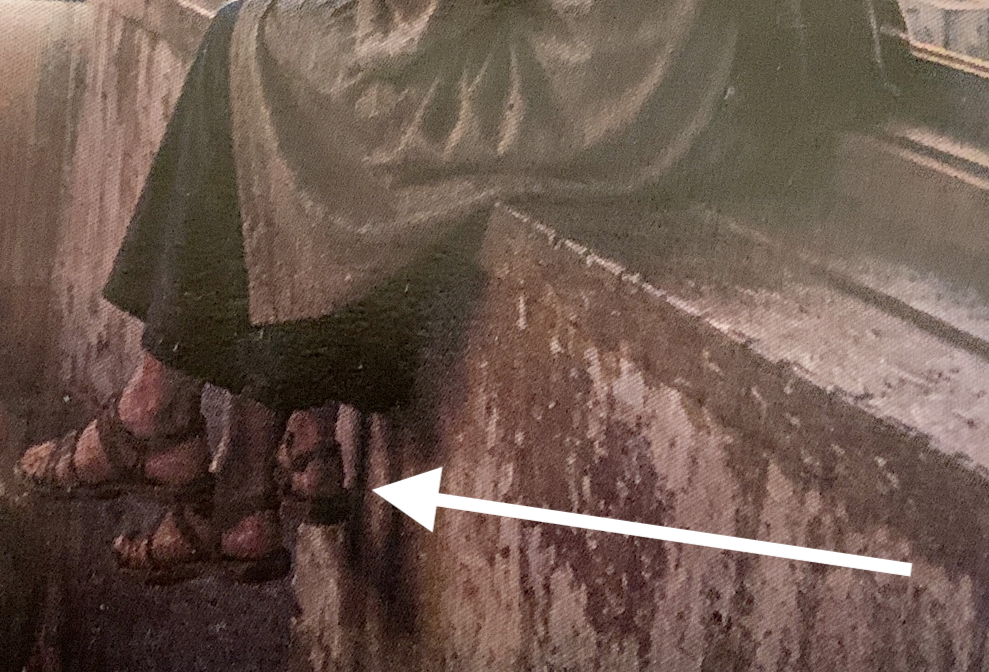 Person in a robe sitting on a stone ledge with several pairs of sandals lined along the wall.